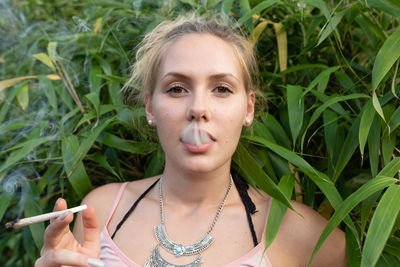 Portrait of young woman smoking marijuana joint while standing against plants