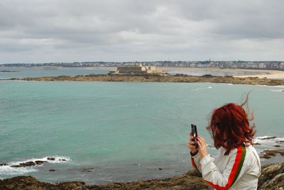 A woman with a cell phone in front of the beach with an island in the background.