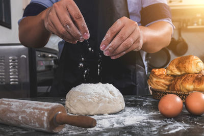 Midsection of woman sprinkling flour on dough in kitchen