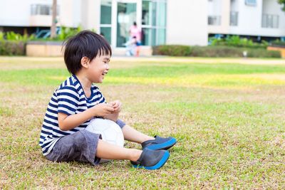 Asian little boy sitting alone with a soccer ball on the lawn in the public park.