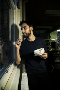 Young man having french fries while standing by window sill