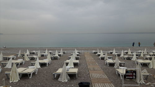 Deck chairs with parasols arranged at beach against cloudy sky