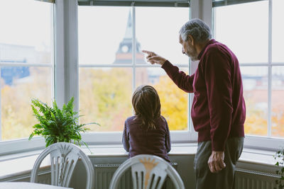 Side view of senior man showing something to great grandson through window at home