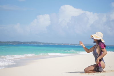 Side view of woman kneeling at beach against cloudy sky