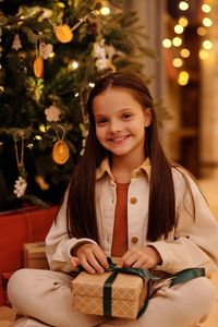Portrait of a smiling young woman sitting on illuminated christmas tree