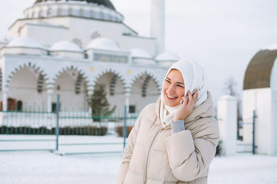 Beautiful smiling young muslim woman in headscarf using mobile against mosque in winter season