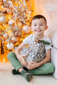 Happy boy sits on floor with gift in his hand by window in front christmas tree