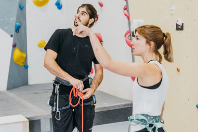 Attentive sportspeople with belay in activewear pointing and looking up while standing near artificial climbing wall during training in gym