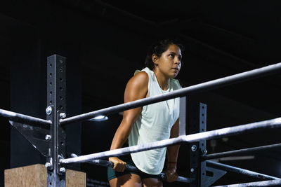 Strong hispanic female athlete in sportswear doing exercise on metal crossbar during intense workout in light gym
