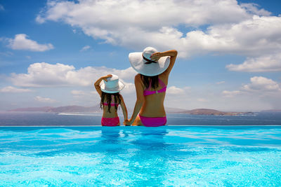Rear view of mother and daughter in bikini sitting by swimming pool while looking at sea