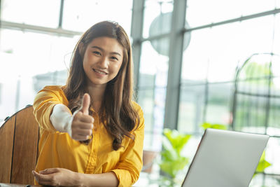 Portrait of smiling businesswoman showing thumbs up while sitting in office