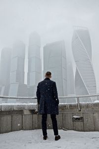 Rear view of man standing against skyscrapers during snowfall