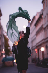 Beautiful woman moving green scarf while holding illuminated jar in city at dusk