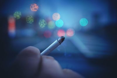 Close-up of cropped hand holding burning cigarette by defocused lights