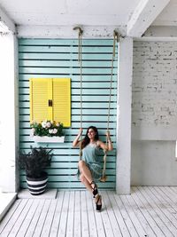 Full length of young woman sitting on swing against wall