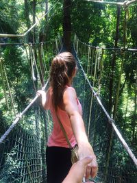 Woman holding cropped hand while standing on footbridge in forest