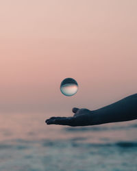 Cropped hand throwing crystal ball against sky during sunset