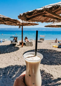 Personal perspective of person holding glass of cold iced coffee on beach in summer.