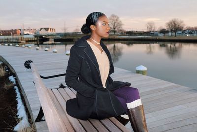 Thoughtful young woman sitting on bench by lake against sky during sunset