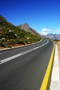 Empty road leading towards mountains against blue sky during sunny day