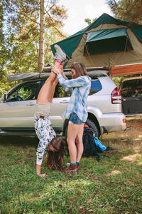 Woman helping friend in doing handstand by car at forest