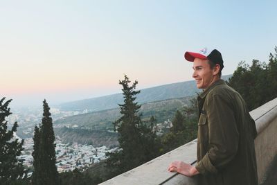 Side view of smiling man standing by concrete wall against sky