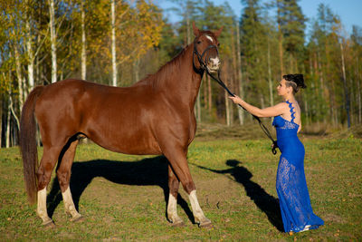 Side view full length of woman standing by horse at forest