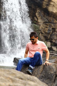 A young man wearing goggles sitting by a waterfall