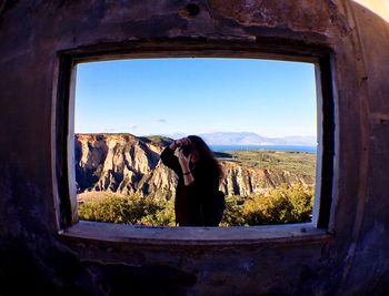 Woman photographing from camera against mountain seen through window on weathered wall