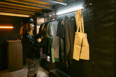 Side view of serious female standing near metal rack with various t shirts while choosing clothes in store with dim light