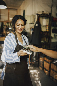 Smiling female owner receiving payment from customer via tap to pay method at workshop