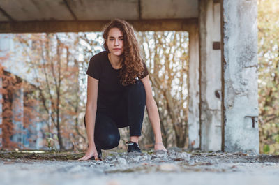 Curly-haired teenage girl in black jeans squats on abandoned construction. vacation outdoor.
