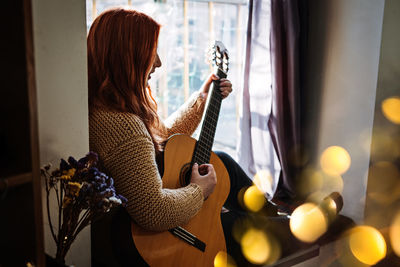 Unaltered candid portrait of young red haired woman in sweater playing acoustic guitar sitting by