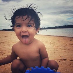 Shirtless baby girl sticking out tongue while playing at beach against sky