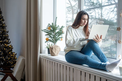 Woman in warm white winter sweater sitting at windowsill at home at christmas eve working on laptop