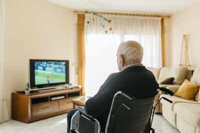 Back view of senior man sitting in wheelchair watching tv at home