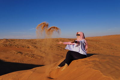 Full length of happy woman wearing hijab throwing sand on desert against sky