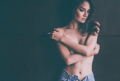 Shirtless woman holding cigarette while standing against wall