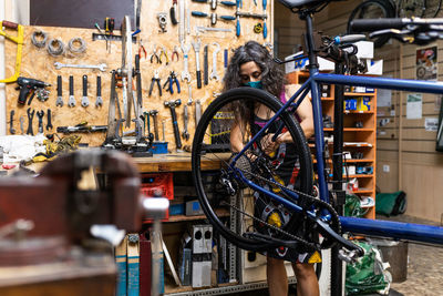 Side view of happy woman in workwear and protective face mask repairing wheel of bike during maintenance service in workshop