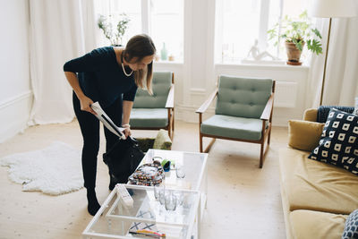 Full length of mid adult woman putting documents in bag while standing in living room at home