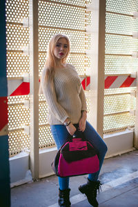 Portrait of young woman holding bag while standing by metal wall