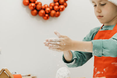 A boy in a santa hat prepares christmas cookies in the kitchen with a christmas wreath on the wall.