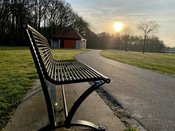 Empty bench in park by building against sky during sunset