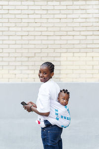 Happy mother holding mobile phone carrying daughter in baby carrier by wall