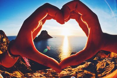 Close-up of hand holding heart shape against sea during sunset