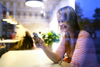 Happy woman using phone in illuminated cafe seen through window