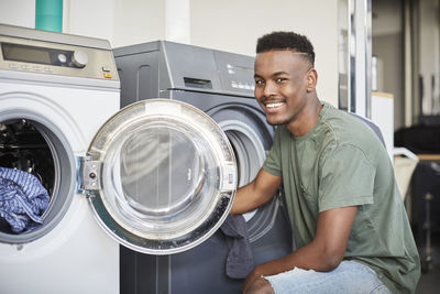 Portrait of smiling young man kneeling by washing machine at laundromat