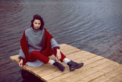 Woman in a grey sweater and a red knitted scarf sits on a bridge in autumn