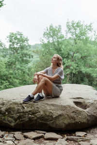 Young woman in summer dress sitting on a big rock in the forest, having rest or meditating