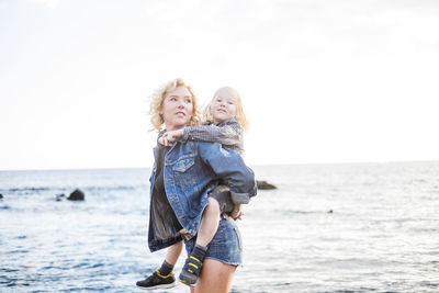 Mother piggybacking cute son while walking at beach against sky during sunset